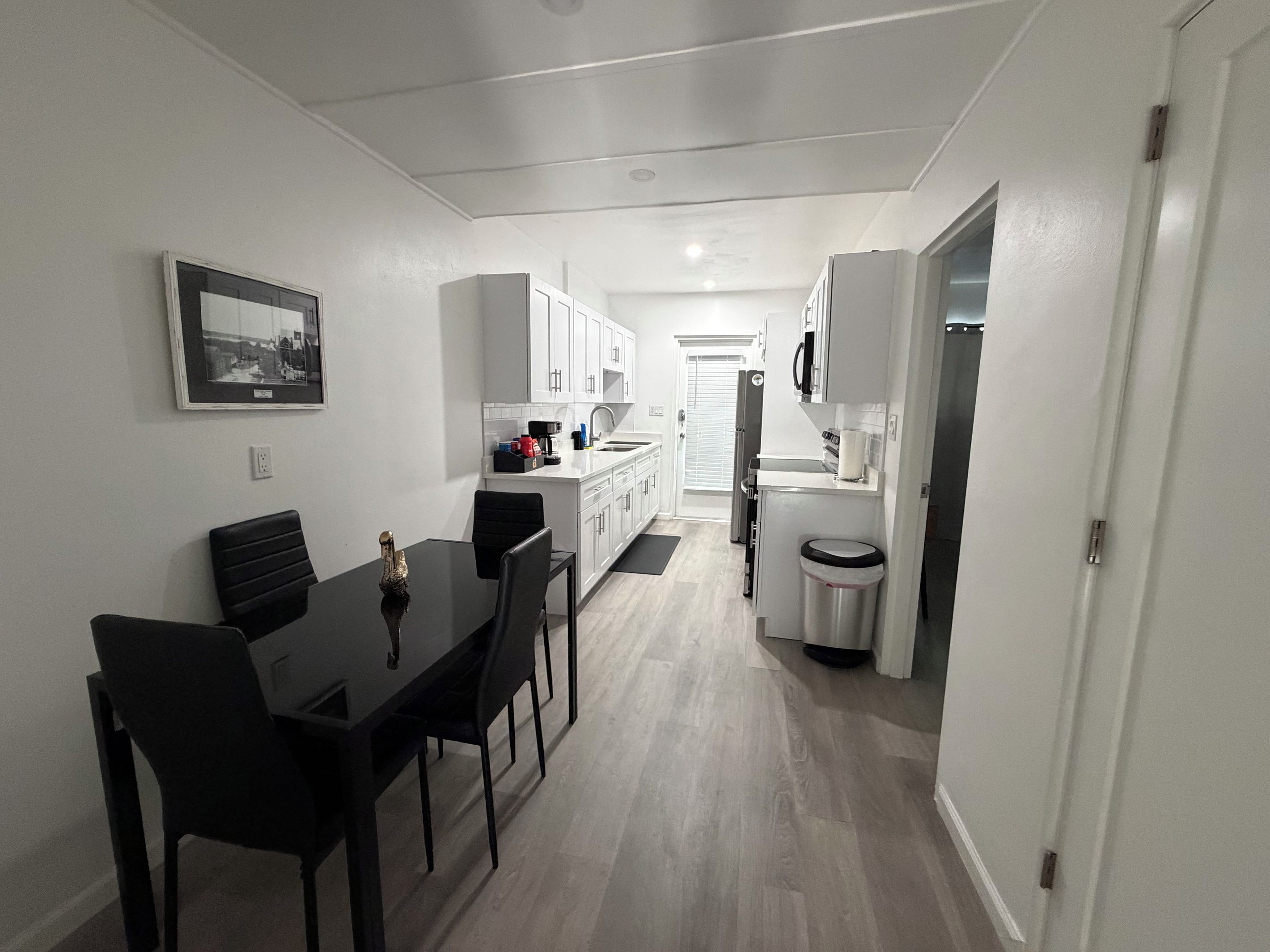 A compact kitchen with white cabinets, a dining area with a black table and chairs, and a small trash bin.