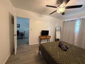 A bedroom with a bed, a ceiling fan, a TV on a stand, a mirror, and a door leading to a dining area.
