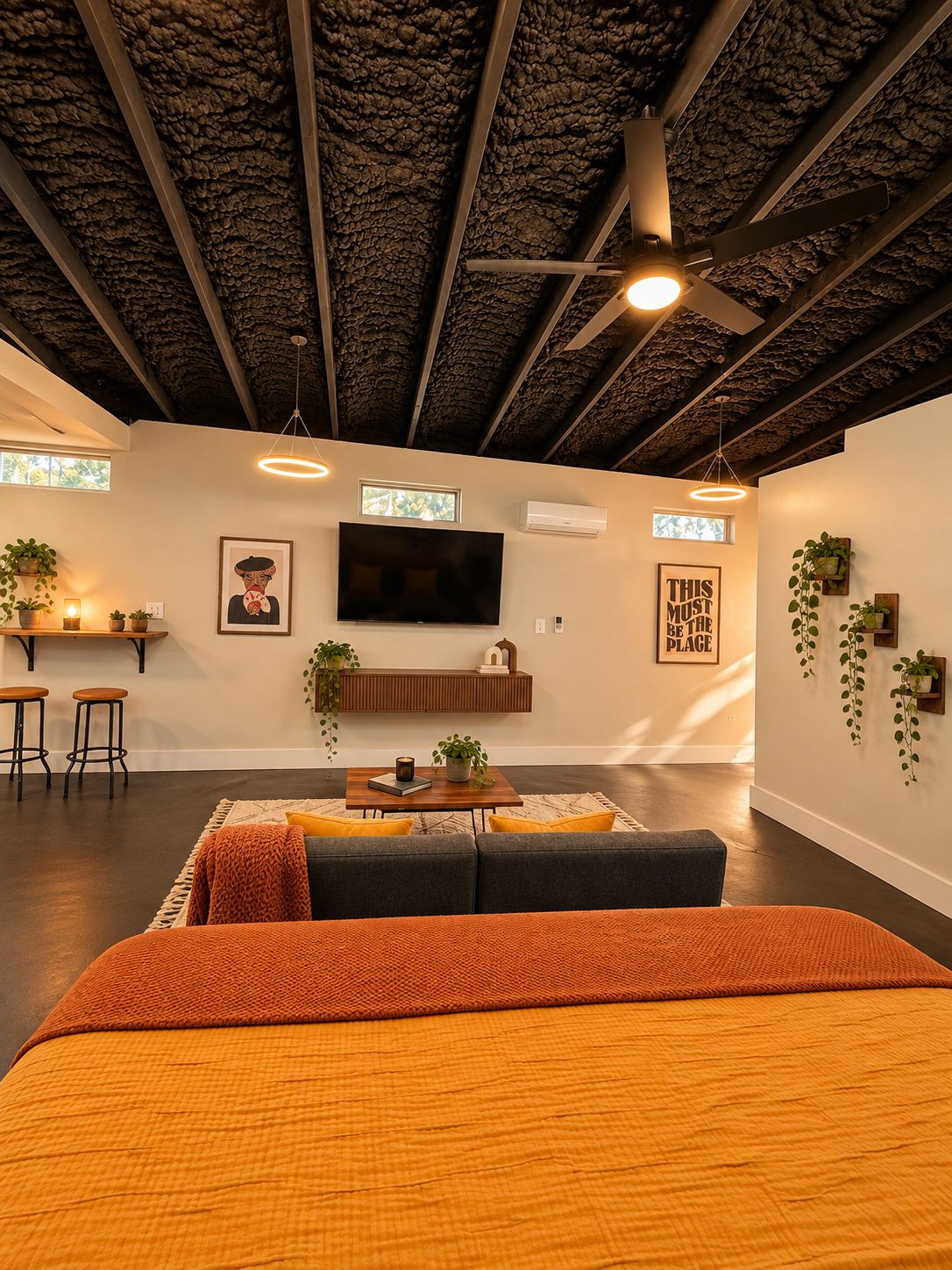 A modern living room with a sofa, a coffee table, and a TV mounted on the wall. There are plants on the windowsill and a ceiling fan.