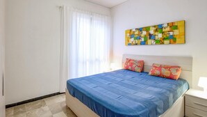 Ein Schlafzimmer mit einem Bett, einem Nachttisch, einer Lampe, einem Fenster mit Vorhängen und einem farbenfrohen abstrakten Gemälde an der Wand.