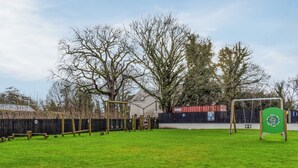 A grassy playground with swings and a climbing structure, surrounded by trees and a fence. - 3 bedroom accommodation in Carnforth (Carnforth)