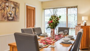 A dining room with a wooden table set for six, surrounded by chairs. There is a vase with flowers on the table, and a window with blinds in the background. - 3 bedroom accommodation in Carnforth (Carnforth)
