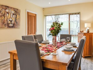 Ein Esszimmer mit einem Holztisch, gedeckt für sechs Personen, umgeben von Stühlen. Auf dem Tisch steht eine Vase mit Blumen, im Hintergrund ist ein Fenster mit Jalousien zu sehen.