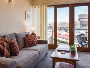 A living room with a sofa, a table with a laptop and a plant, a lamp, and a view of a river and buildings through a sliding door. - 3 bedroom accommodation in Carnforth (Carnforth)