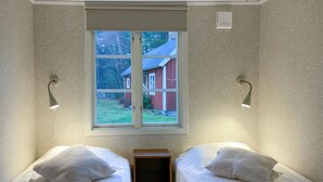 Une petite chambre accueillante avec deux lits simples, une fenêtre donnant sur une maison rouge et des lampes de lecture fixées au mur.