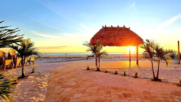 A beach with a thatched-roof structure, palm trees, and a paved walkway. - Casa Playa #403 Beach Front - Dolphin Watch (Fort Myers Beach)