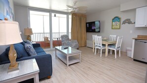 A living room with a sofa, coffee table, and dining area with chairs. There is a television mounted on the wall and a ceiling fan. - Casa Playa #403 Beach Front - Dolphin Watch (Fort Myers Beach)