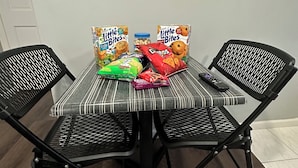 A dining table with a striped tablecloth, two black chairs, and various snack packages including Little Bites cereal and chips. - Charming 2-bedroom apartment in wonderful Queens with WiFi and Parking (Queens)