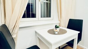 Una habitación con una ventana blanca, una pequeña mesa blanca con una planta y una silla negra.