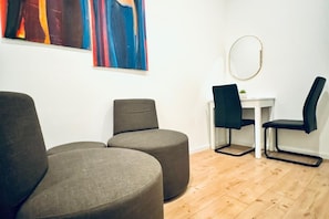 A room with a wooden floor, two grey armchairs, a small white table with a chair, and a round mirror on the wall.
