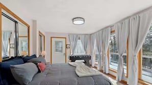 A spacious bedroom with a large bed, a desk, and a view of the outdoors through large windows. - Pikes Peak Views & Hot Tub | Mountain Home (Woodland Park)