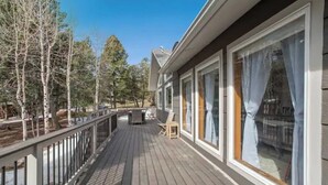 A wooden deck with a railing, large windows with curtains, and a view of trees and a building in the background. - Pikes Peak Views & Hot Tub | Mountain Home (Woodland Park)