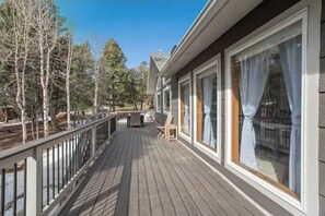 A wooden deck with a railing, large windows with curtains, and a view of trees and a building in the background.
