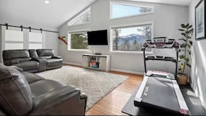 A modern living room with a sectional sofa, a treadmill, and large windows offering a view of the outdoors. - Pikes Peak Views & Hot Tub | Mountain Home (Woodland Park)