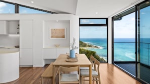 A modern kitchen with white cabinetry, a wooden dining table with four chairs, and a view of the ocean through large windows. - Iluka Lookout | Unforgettable ocean views, new home (Wye River)