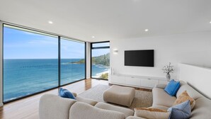 A modern living room with a large window offering a view of the ocean and a distant coastline. - Iluka Lookout | Unforgettable ocean views, new home (Wye River)