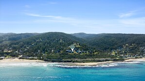 A coastal area with a beach, forested hills, and a cluster of buildings. - Iluka Lookout | Unforgettable ocean views, new home (Wye River)