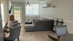 Una sala moderna con un sofá gris, una mesa de comedor con sillas y una cocina visible a través de una puerta abierta.