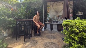 Een man zit op een krukje onder een pergola in een tuin met potplanten en een betegelde vloer.