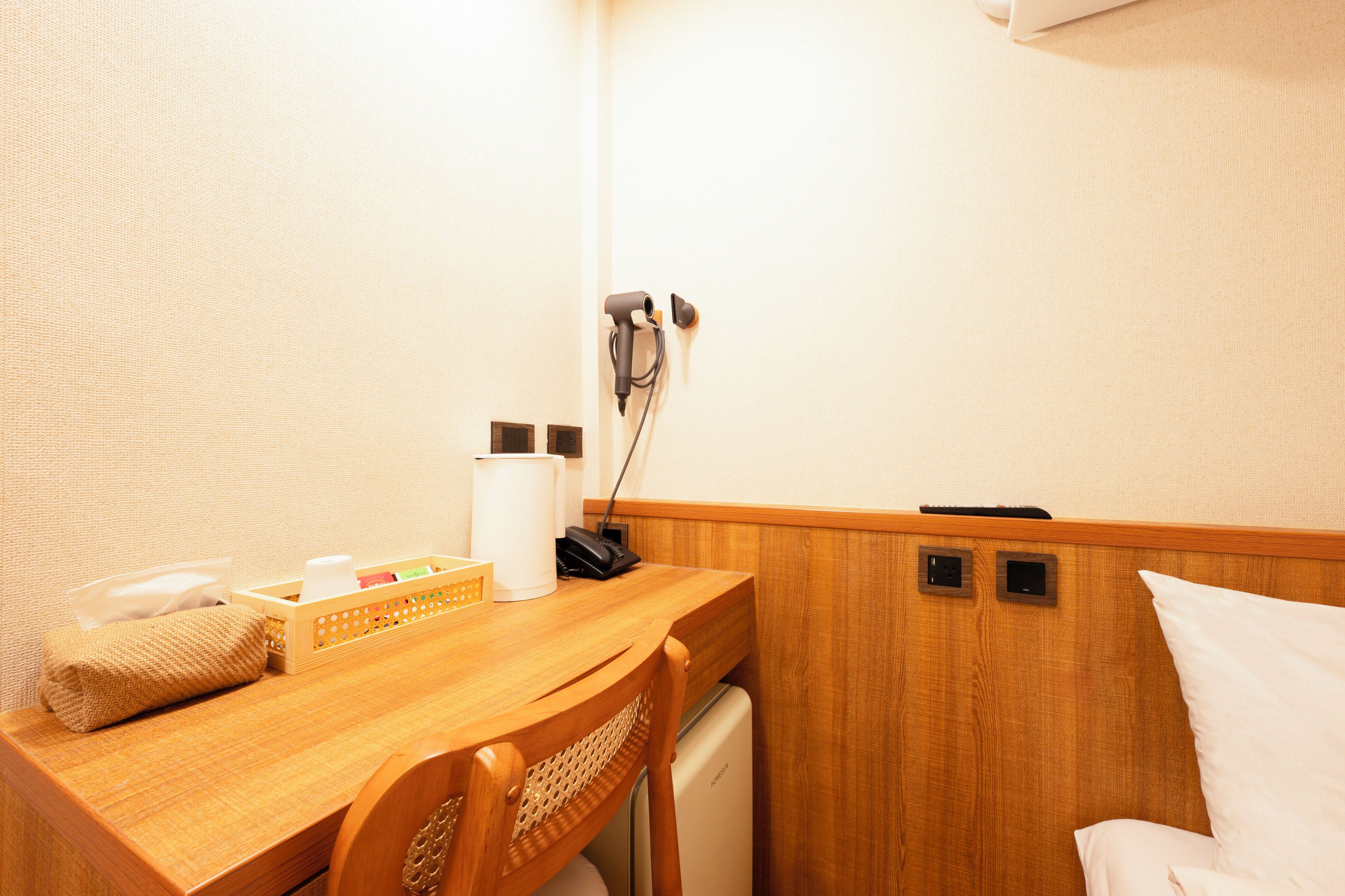 A small, compact hotel room with a wooden desk, a chair, a bed, and a small kitchenette area.