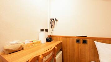 A small, compact hotel room with a wooden desk, a chair, a bed, and a small kitchenette area.