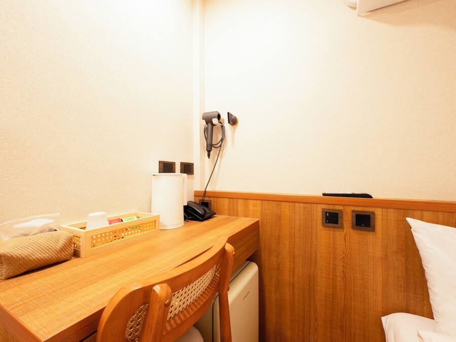 A small, compact hotel room with a wooden desk, a chair, a bed, and a small kitchenette area.