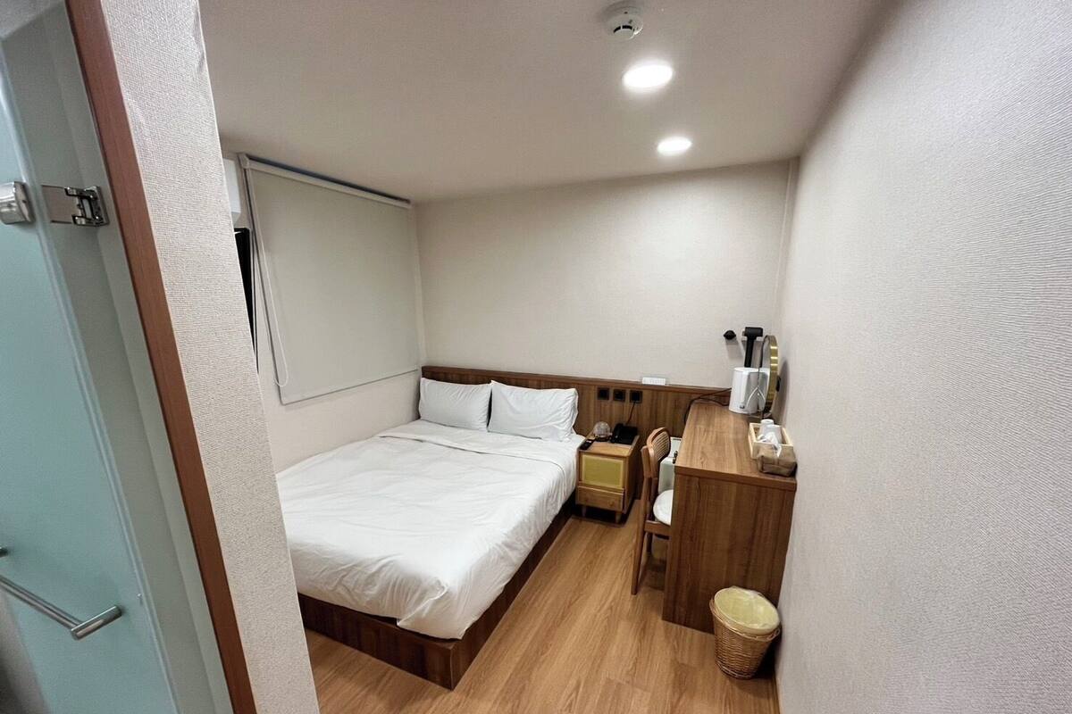 A hotel room with a single bed, a wooden desk, a chair, a bedside table, a waste basket, and a ceiling light.