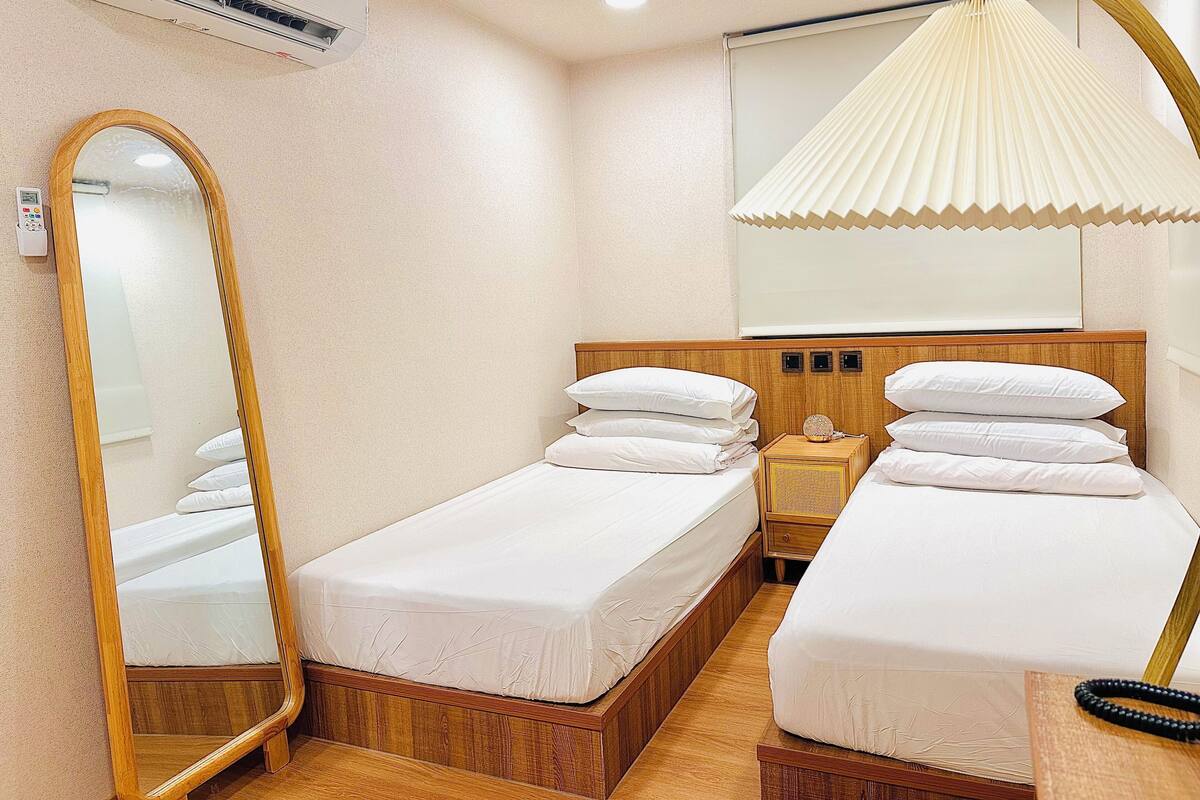 A hotel room with two single beds, a wooden headboard, a bedside table with a lamp, a mirror, and an air conditioning unit.