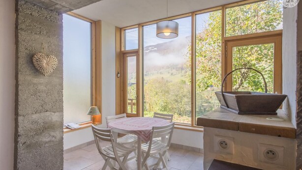 A cozy room with a large window, a round table with chairs, a wooden desk, and a hanging light fixture.