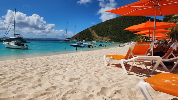 A beach with lounge chairs and orange umbrellas, boats in the water, and a hilly background. - MUST SELL 1-3 wks ALL INCLUSIVE crewed BVI private yacht charter FP 67 Catamaran (Tortola)