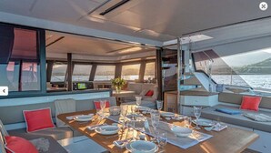 A yacht's dining area with a table set for four, surrounded by comfortable seating and large windows offering a view of the sea. - MUST SELL 1-3 wks ALL INCLUSIVE crewed BVI private yacht charter FP 67 Catamaran (Tortola)