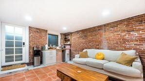 Un espacio habitable compacto con un sofá blanco, una mesa de centro de madera, una zona de cocina con refrigerador y microondas, y una pared de ladrillo.