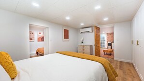Un dormitorio con una cama grande, un aparador y una zona de escritorio con una silla. Se ve una puerta que conduce a otra habitación con pared de ladrillo y televisor.