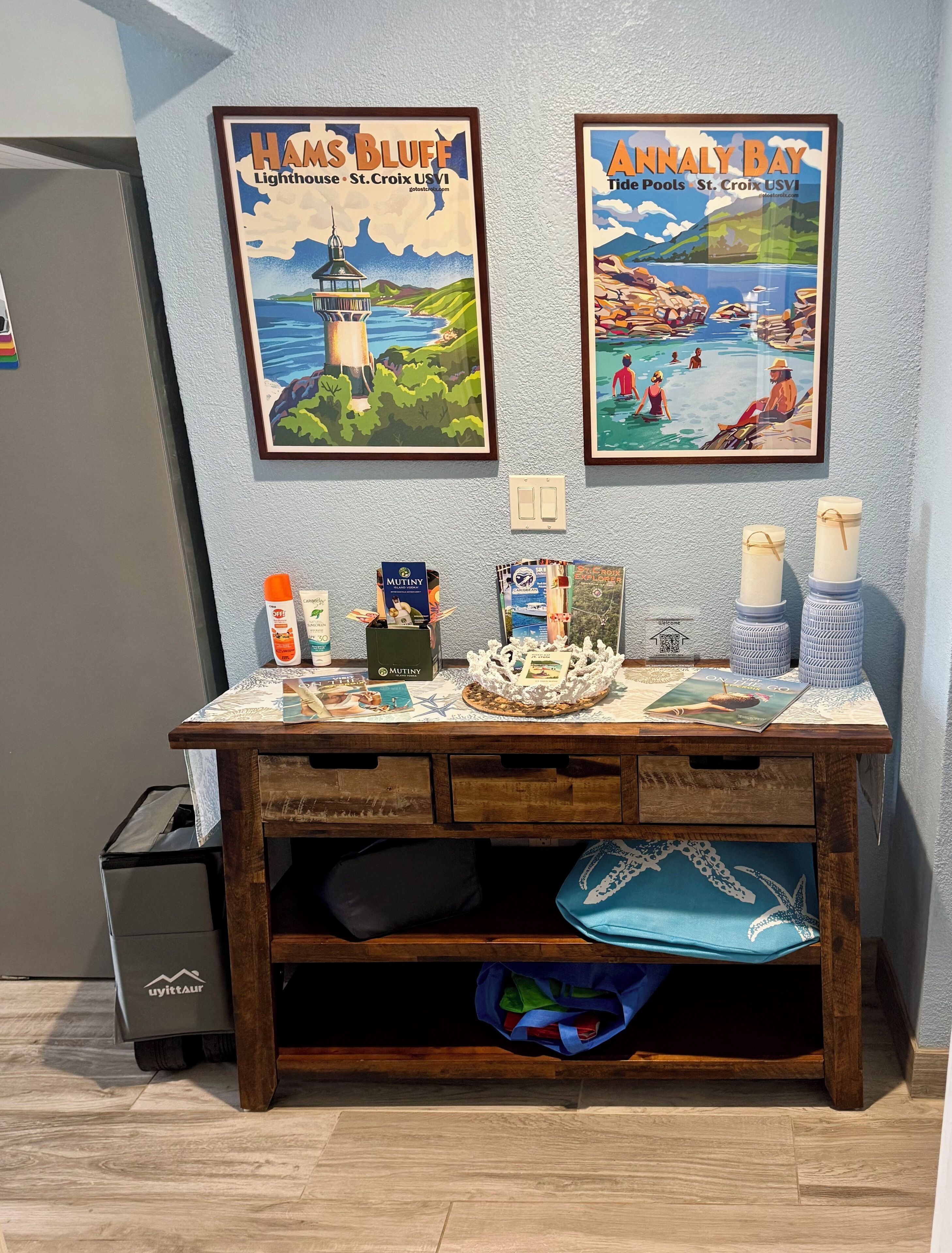 A wooden desk with framed posters of 'Haas Bluff Lighthouse' and 'Annaly Bay Tide Pools'. The desk has various items like a Mutiny box, a candle, and a blue bag.