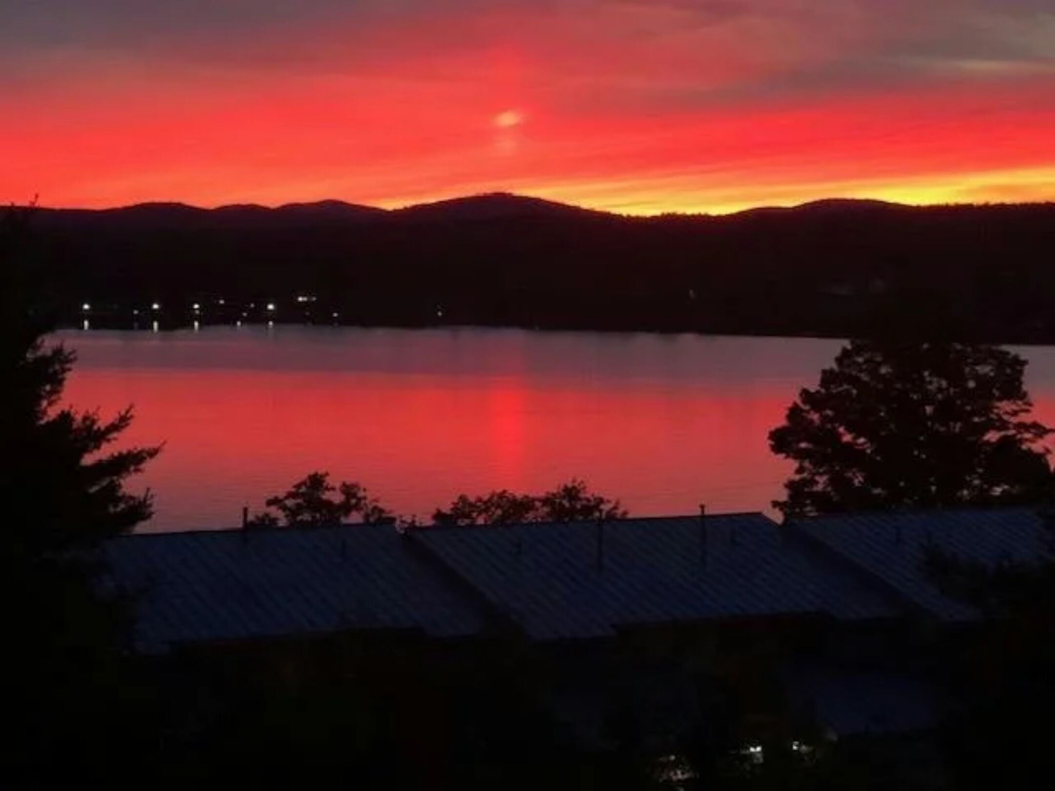 A sunset over a calm lake with a silhouette of a mountain range in the background and a building with a sloped roof in the foreground.
