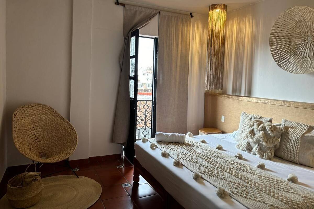 A bedroom with a bed, a wicker chair, a window with curtains, and a wall-mounted lamp.