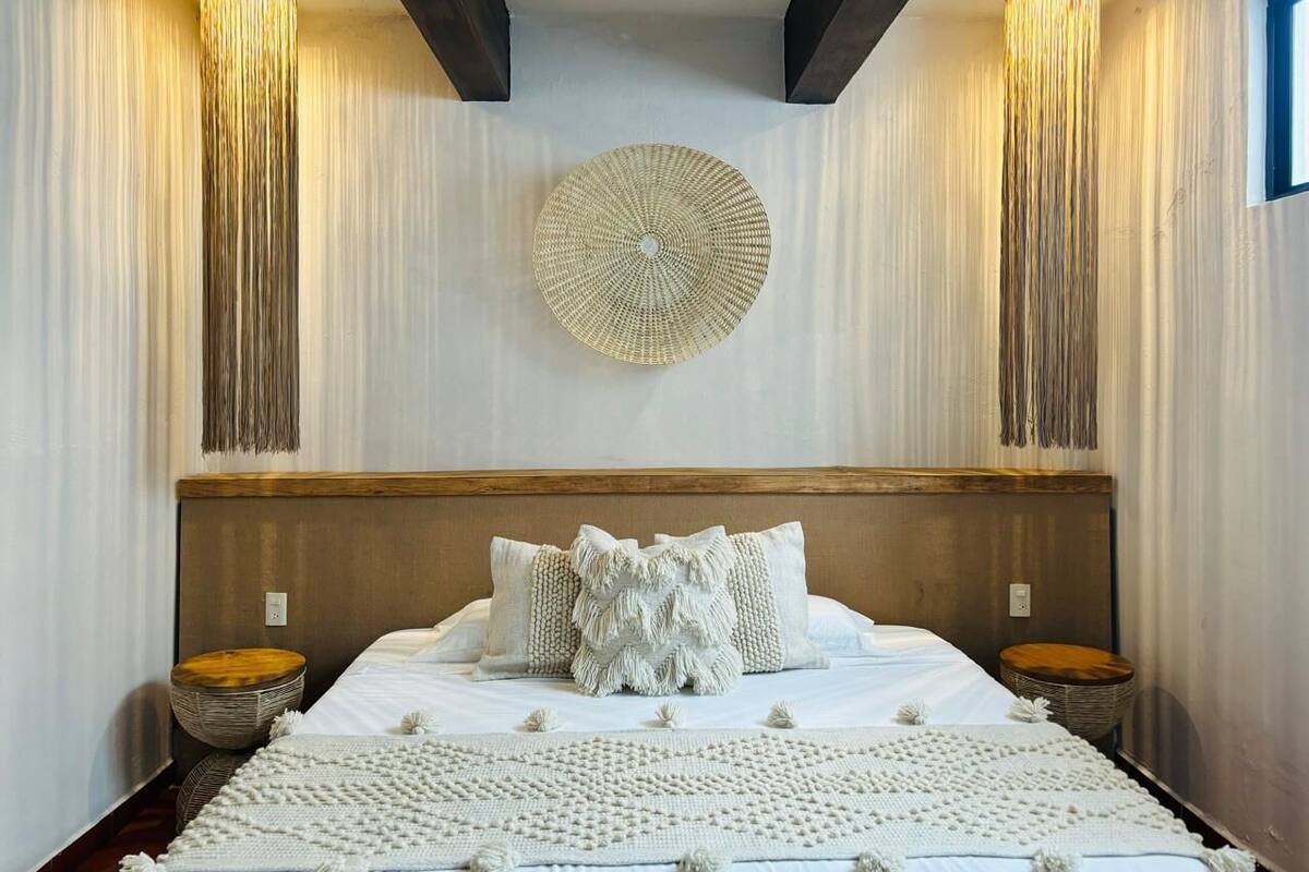 A bedroom with a large bed, two nightstands, a woven wall hanging, and vertical light fixtures.