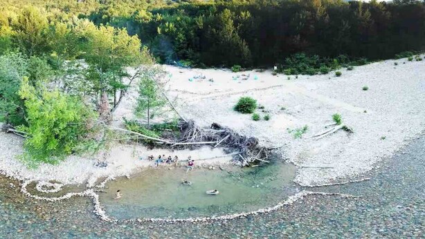 Ein Flussufer mit einer kleinen Brücke, Bäumen und einem Berg im Hintergrund.