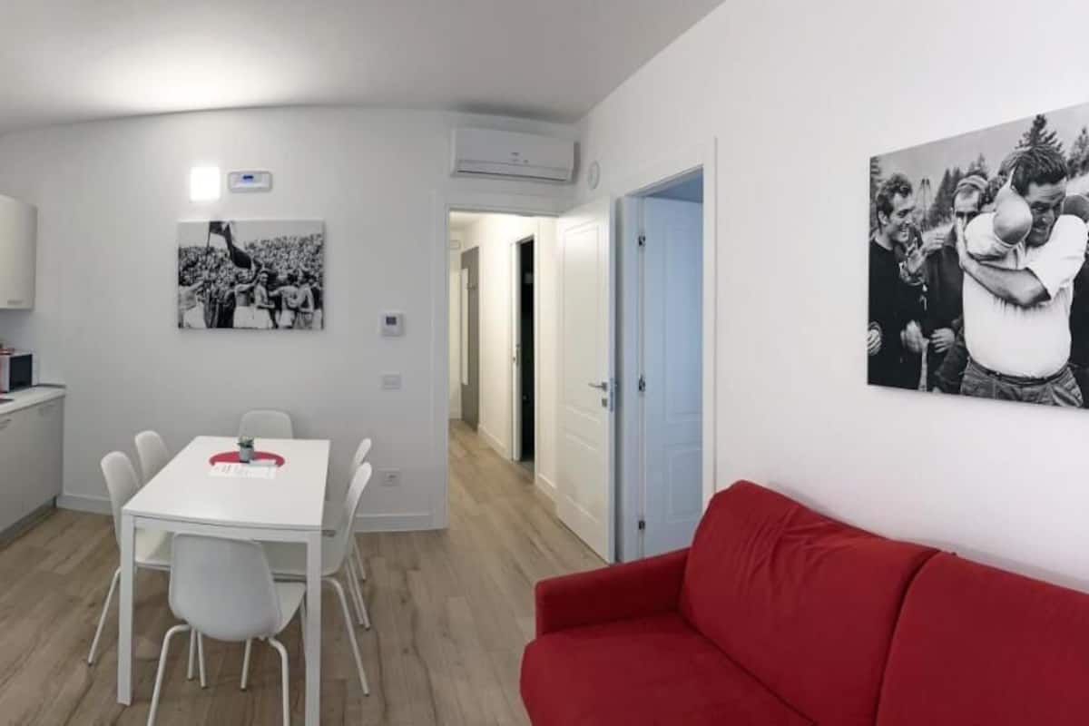 A compact room with a bed, a dining table with chairs, a red sofa, a kitchen area, and two wall-mounted air conditioning units.
