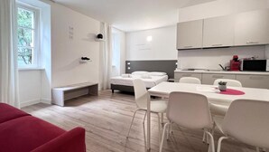 A modern, minimalist hotel room with a bed, a small desk, a dining table with chairs, and a kitchenette. - City Gallery Apartments (Trieste)