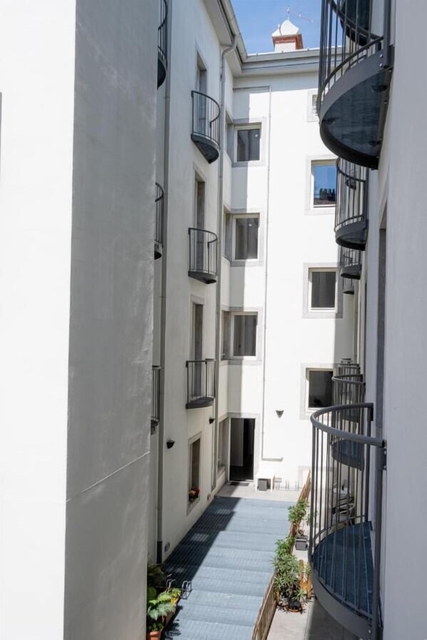 A narrow corridor between two white buildings with balconies, a small outdoor area with plants, and a clear sky.