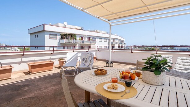 En tagterrasse med et bord dækket til morgenmad, plantekasser og udsigt til en hvid bygning over vandet.