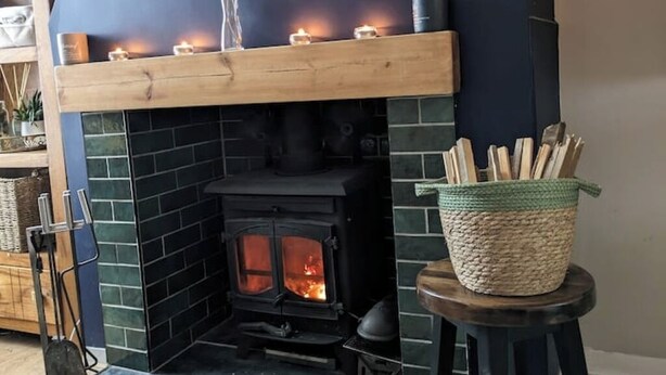 Una estufa de leña con un fuste de madera encima, velas encendidas y una cesta tejida sobre un taburete al lado de la estufa.