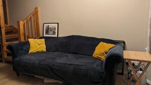 Un sofá de color oscuro con cojines amarillos, una silla de madera y una pequeña mesa de madera en una habitación con paredes lisas.