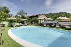 Una piscina con sillas de descanso y sombrillas, rodeada de follaje y un edificio de piedra.