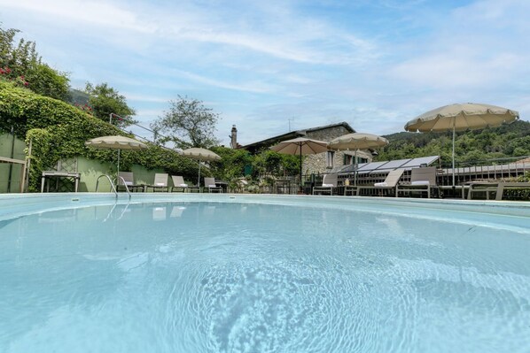 Une piscine entourée de verdure et de bâtiments, avec des chaises longues et des parasols.