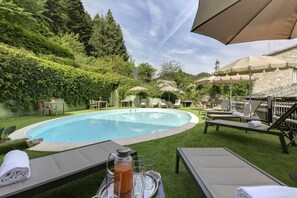 A swimming pool surrounded by lounge chairs and umbrellas, with a stone wall and greenery in the background.