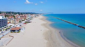 A coastal cityscape with a wide sandy beach, a clear blue sea, and a long pier extending into the water.