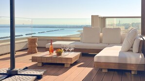 A rooftop terrace with a view of the sea, a wooden coffee table with a bottle and glasses, and a white sofa.