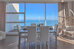 Uma sala de jantar com vista para o mar, um edifício alto e uma cadeira de vime.